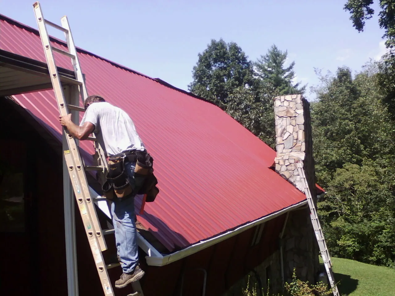 Roofer on a ladder beside a red metal roof and stone chimney — Soffit & Fascia Repair by Roofing Brothers in Cedarpines Park